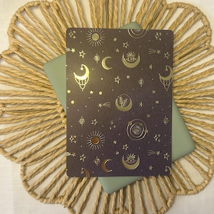 Pode incluir: Um cartão roxo escuro com desenhos celestiais dourados, incluindo luas, estrelas e sóis, repousa sobre uma superfície verde clara. O cartão é colocado sobre um tapete de mesa bege tecido com um design floral, criando uma estética mística.