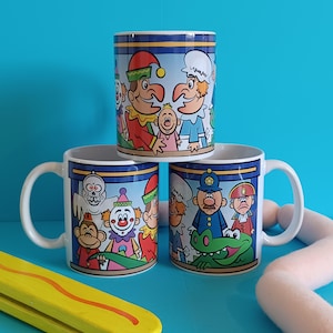 May include: Three ceramic mugs with colourful cartoon illustrations. The mugs feature characters with different expressions and poses. The mugs are white with a blue rim and handle.