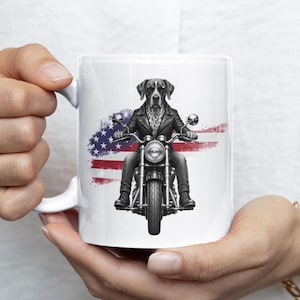 May include: A white ceramic mug featuring a black and white illustration of a dog wearing a leather jacket and sunglasses while riding a motorcycle. Behind the dog is a stylised, distressed graphic of the American flag in red, white, and blue.