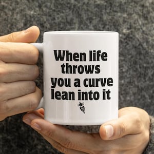 May include: White ceramic mug with the text "When life throws you a curve lean into it" in black. A small graphic of a motorcycle is below the text. The mug is held by a person.