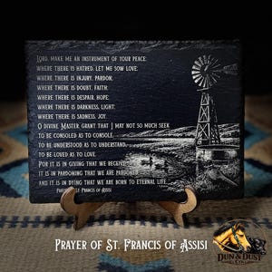 May include: Black slate plaque with the "Prayer of St. Francis of Assisi" engraved in white. The design includes a windmill and a lake scene. The plaque is displayed on a wooden stand.