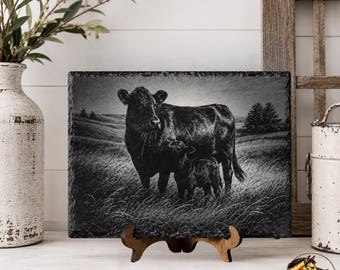 May include: A rectangular black slate plaque featuring a laser-engraved image of a cow and a calf standing in a grassy field. The plaque has a rough, natural stone edge and is displayed on a small wooden stand. The engraving shows detailed textures.