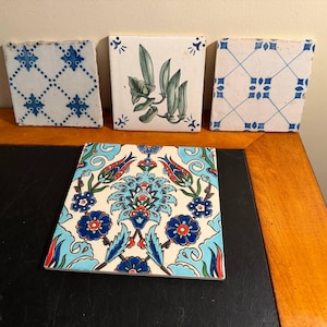 May include: Four square ceramic tiles. Three are upright, featuring blue lattice patterns and a green pea pod illustration. The fourth tile lies flat, displaying a vibrant, symmetrical floral pattern in shades of blue, red, and green on a white background.