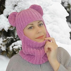 May include: A pink knitted balaclava with ear flaps in the shape of bear ears. The balaclava is made of a soft, fuzzy yarn.