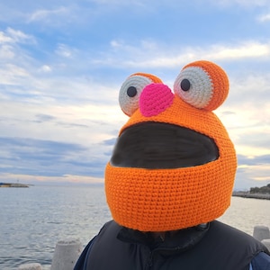 May include: Orange crocheted motorcycle helmet with large white eyes, a pink nose, and a black visor.