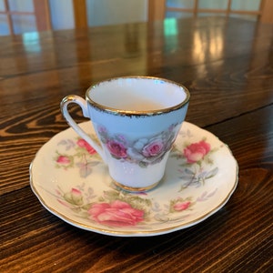 Op de afbeelding: Een delicate kop en schotel set. De witte porseleinen kop en schotel zijn versierd met roze rozen en groene bladeren. Gouden randen accentueren de randen van de kop en schotel, wat een vleugje elegantie toevoegt. De set staat op een donkere houten tafel.