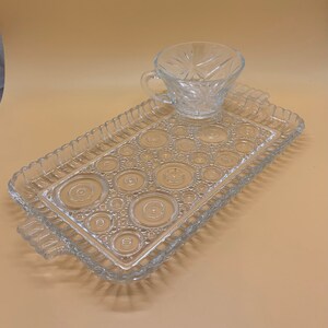 Puede incluir: Juego de taza de té y bandeja de servir de vidrio transparente. La taza de té tiene un diseño de estrella. La bandeja rectangular presenta un patrón de círculos y un borde con cuentas. El juego es de vidrio transparente.