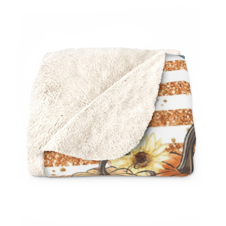 May include: A soft, plush throw blanket with a white and orange striped pattern. The blanket features a graphic of a pumpkin with sunflowers and glitter accents.