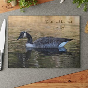 May include: Rectangular cutting board featuring a serene image of a goose swimming in water. The goose has black, white, and gray feathers. The text "Be still and know that I am God" is printed on the board, along with the reference Psalms 46:10.