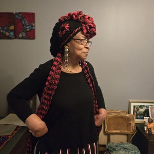 May include: A woman wearing a black long-sleeve shirt, a black and red patterned scarf, and a black and red crocheted hat. She is also wearing glasses and large earrings. The background includes artwork and a television.