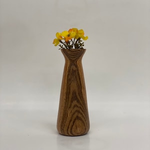 Puede incluir: Un jarrón de madera con un patrón de veta de madera marrón y marrón oscuro. El jarrón es estrecho en la parte superior y se ensancha hacia la parte inferior. Está lleno de un pequeño ramo de flores amarillas.
