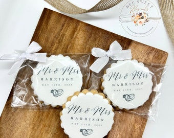 May include: Three white round cookies with scalloped edges, decorated with black text and a small illustration of two rings. The cookies are individually packaged in clear cellophane bags with white ribbon bows. The text on the cookies reads "Mr & Mrs Harrison May 11th, 2023".