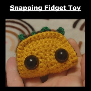 Könnte beinhalten: Ein gehäkeltes Taco-förmiges Fidget-Spielzeug mit Glitz Augen. Der Taco ist gelb mit grünem Salat und braunem Fleisch. Es ist eine lustige und einzigartige Möglichkeit, Stress abzubauen.