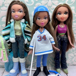 Peut inclure: Trois poupées mannequins aux longs cheveux bruns. La poupée de gauche porte une veste bleu clair, un haut vert, un jean foncé et des bottes bleu clair. La poupée centrale porte un casque bleu, un maillot blanc avec le numéro 34 et des patins. La poupée de droite porte un haut rose et un jean foncé.