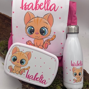 Può includere: Uno zaino rosa con un pannello frontale bianco che presenta un gatto dei cartoni animati e il nome "Isabella". Sono inclusi anche una lunchbox e una borraccia abbinate con lo stesso design.