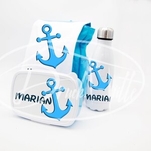 Può includere: Una borsa per il pranzo bianca con un design a forma di ancora blu, una scatola per il pranzo bianca con un design a forma di ancora blu e il nome "MARIAN" su di essa, e una bottiglia d'acqua bianca con un design a forma di ancora blu e il nome "MARIAN" su di essa.