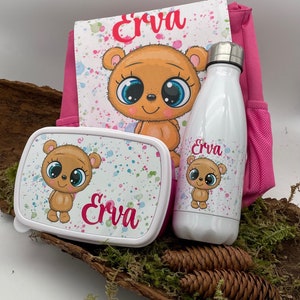 Può includere: Zaino rosa con una borsa pranzo bianca e una borraccia bianca. Tutti e tre gli articoli presentano un orso dei cartoni animati con occhi grandi e il nome "Erva" stampato su di essi.