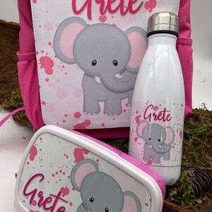 Può includere: Zaino rosa, borsa pranzo e borraccia con sfondo bianco e design a spruzzo rosa. Ogni articolo presenta un elefante grigio con orecchie rosa e il nome "Grete" in lettere rosa.