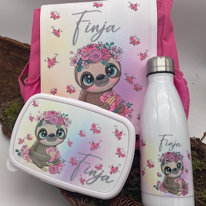 Può includere: Uno zaino rosa con un designo di bradipo bianco, una scatola per il pranzo bianca con un designo di bradipo e una bottiglia d'acqua bianca con un designo di bradipo. Tutti e tre gli articoli hanno il nome "Finja" stampato su di essi.