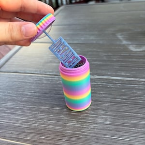 May include: A rainbow-striped bubble wand container with a matching lid. The lid is being held open, revealing the bubble wand. The wand has the text "DEAR WIFE" on it. The container is cylindrical.