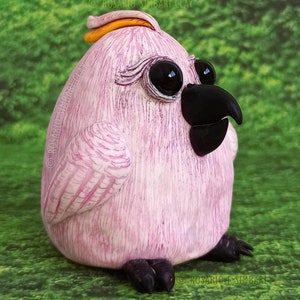 Puede incluir: Una figurita de pájaro rosa con plumas, ojos negros y pico negro. El pájaro tiene una pequeña cresta naranja en la cabeza y está sentado sobre una superficie verde cubierta de hierba.