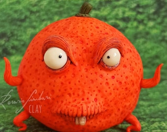 Puede incluir: Una escultura redonda de arcilla de color naranja brillante que representa a un monstruo con una sonrisa de dientes, ojos saltones y una superficie texturizada. El monstruo tiene dos pequeños brazos curvados y un pequeño tallo verde en la parte superior.