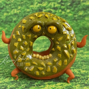 Puede incluir: Una dona verde y naranja con cara y piernas. La dona está cubierta de chispas verdes y amarillas. La dona está de pie sobre una superficie verde cubierta de hierba.