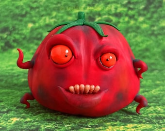 Puede incluir: Una escultura caprichosa de tomate rojo con una expresión traviesa. El tomate tiene ojos anaranjados, una sonrisa con dientes y pequeños brazos y piernas rizados. La escultura está sobre un fondo verde y herboso.