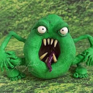 Puede incluir: Escultura de arcilla verde de un monstruo de dibujos animados con la boca muy abierta, dientes afilados y una lengua morada. El monstruo tiene ojos blancos y posa con los brazos y las piernas extendidos. La escultura está sobre un fondo verde.