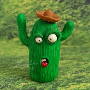 Puede incluir: Figura de cactus verde con sombrero marrón. El cactus tiene grandes ojos blancos, una lengua roja y los brazos levantados. La figura hecha a mano tiene la firma del artista y está sobre un fondo verde.