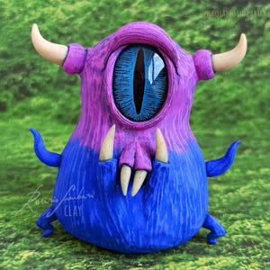 Puede incluir: Una figurita de monstruo hecha a mano de color morado y azul con un solo ojo grande azul y dientes afilados. El monstruo tiene cuernos y una superficie texturizada.