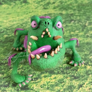 Puede incluir: Una figura de monstruo de arcilla verde con la boca muy abierta, dientes afilados y una larga lengua púrpura. El monstruo tiene ojos rosados, detalles púrpuras y está posado sobre un fondo verde. La firma del artista es visible en la parte inferior izquierda.