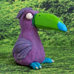Puede incluir: Una caprichosa escultura de pájaro púrpura con un gran pico verde y detalles azules. El pájaro tiene grandes ojos blancos y pies azules. La escultura está sobre un fondo verde. La firma del artista es visible en la parte inferior izquierda.
