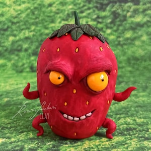 Puede incluir: Una escultura caprichosa de fresa roja con una expresión amenazante. La obra de arte presenta grandes ojos amarillos, una sonrisa con dientes y pequeños brazos y piernas. La fresa tiene semillas amarillas y una parte superior verde y frondosa. La firma del artista es visible en la parte inferior izquierda.