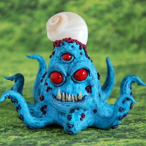 Puede incluir: Un pulpo azul y rojo con tres ojos y una concha en la cabeza. El pulpo tiene una sonrisa de dientes y está hecho de arcilla polimérica.