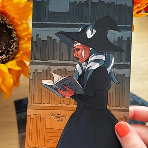 May include: Illustration of a character in a black witch hat and dress, reading a book in front of a bookshelf. The character has white and blue markings on their face and head. The book's title is "Axes & Roses". Signed by Allison Rosemary Watt.