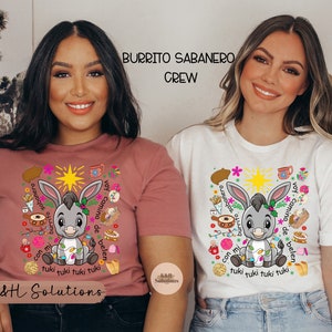 Könnte beinhalten: Zwei Frauen tragen T-Shirts mit einem Cartoon-Esel-Design. Der Esel ist von farbenfrohen Illustrationen und Text umgeben, der "Burrito Sabanero Crew" und "tuki tuki tuki tuki" lautet. Ein Shirt ist staubrosa, das andere ist weiß.