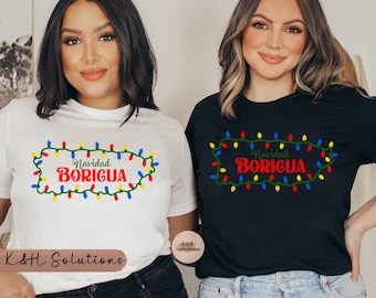 May include: Two women wearing white and black t-shirts with the words "Navidad Boricua" in red, outlined by a green border of Christmas lights. The shirts are a festive design, perfect for the holiday season.
