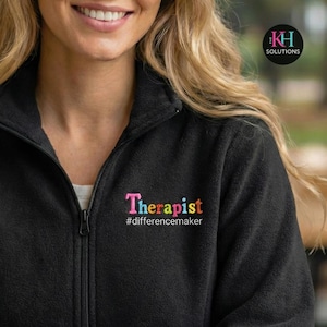 May include: Black fleece zip-up jacket with the word "Therapist" embroidered in pink, yellow, and blue, and the hashtag "#differencemaker". The jacket has a high collar and a silver zipper.