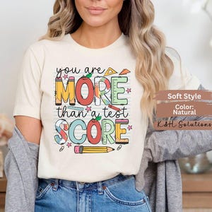 May include: Cream-colored t-shirt with the text "you are MORE than a test SCORE" in colorful, school-themed graphics. The design includes pencils, rulers, and stars. The shirt is a soft style and natural color.