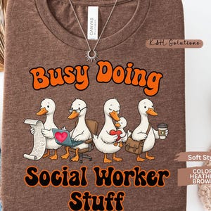 May include: Heather brown t-shirt with the text "Busy Doing Social Worker Stuff" in orange and black. The shirt features a cartoon illustration of four ducks, each engaged in different social worker activities.