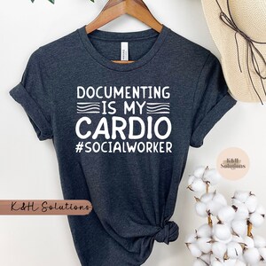 May include: Dark gray t-shirt with the text "DOCUMENTING IS MY CARDIO #SOCIALWORKER" in white. The shirt is hanging on a wooden hanger. The shirt is made of a soft material.