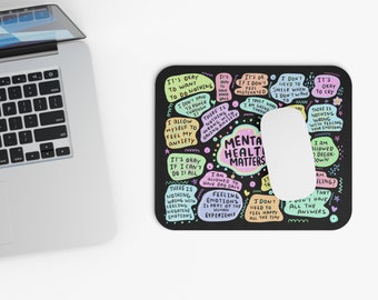 May include: Black mousepad with the text "MENTAL HEALTH MATTERS" and various positive affirmations in colorful speech bubbles. A white computer mouse is on the right side of the mousepad. A laptop is in the upper left corner.
