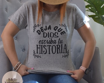 May include: A heather gray t-shirt with the text "Deja que Dios escriba tu HISTORIA" in black script. The shirt is short-sleeved and is being held up by the person wearing it. The person is also wearing a straw hat and blue jeans.