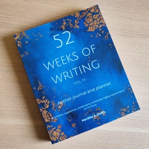 May include: A blue and gold book titled "52 Weeks of Writing Vol. III: Author Journal and Planner". The book cover features a quote from Jamie Sands: "A brilliant, supportive, challenging workbook, highly recommend."