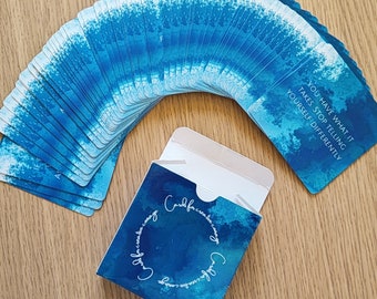 May include: A set of blue and white affirmation cards with a blue and white box. The box has the text "Cards for creative carnage" on it. The cards have affirmations such as "You have what it takes. Stop telling yourself differently."