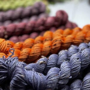 May include: A close-up of a collection of yarn skeins in various colors, including orange, purple, green, and blue. The yarn is arranged in a stack, with the blue yarn at the bottom.