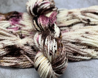 May include: A skein of yarn in a cream, brown, and pink colourway. The yarn is hand-dyed and has a speckled appearance.