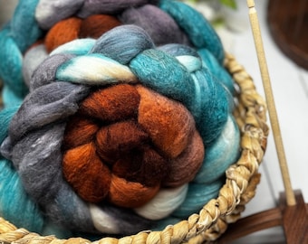 May include: A braided bundle of wool roving in shades of teal, dark gray, white, and burnt orange, resting inside a woven wicker basket. A wooden drop spindle stands upright next to the basket, featuring a thin shaft and a dark wood base.