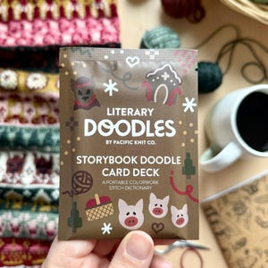May include: A person's hand holding a brown Literary Doodles Storybook Doodle Card Deck. The card deck features illustrations of a gingerbread house, pigs, and snowflakes. The background includes yarn, a mug of coffee, and a knitted swatch.